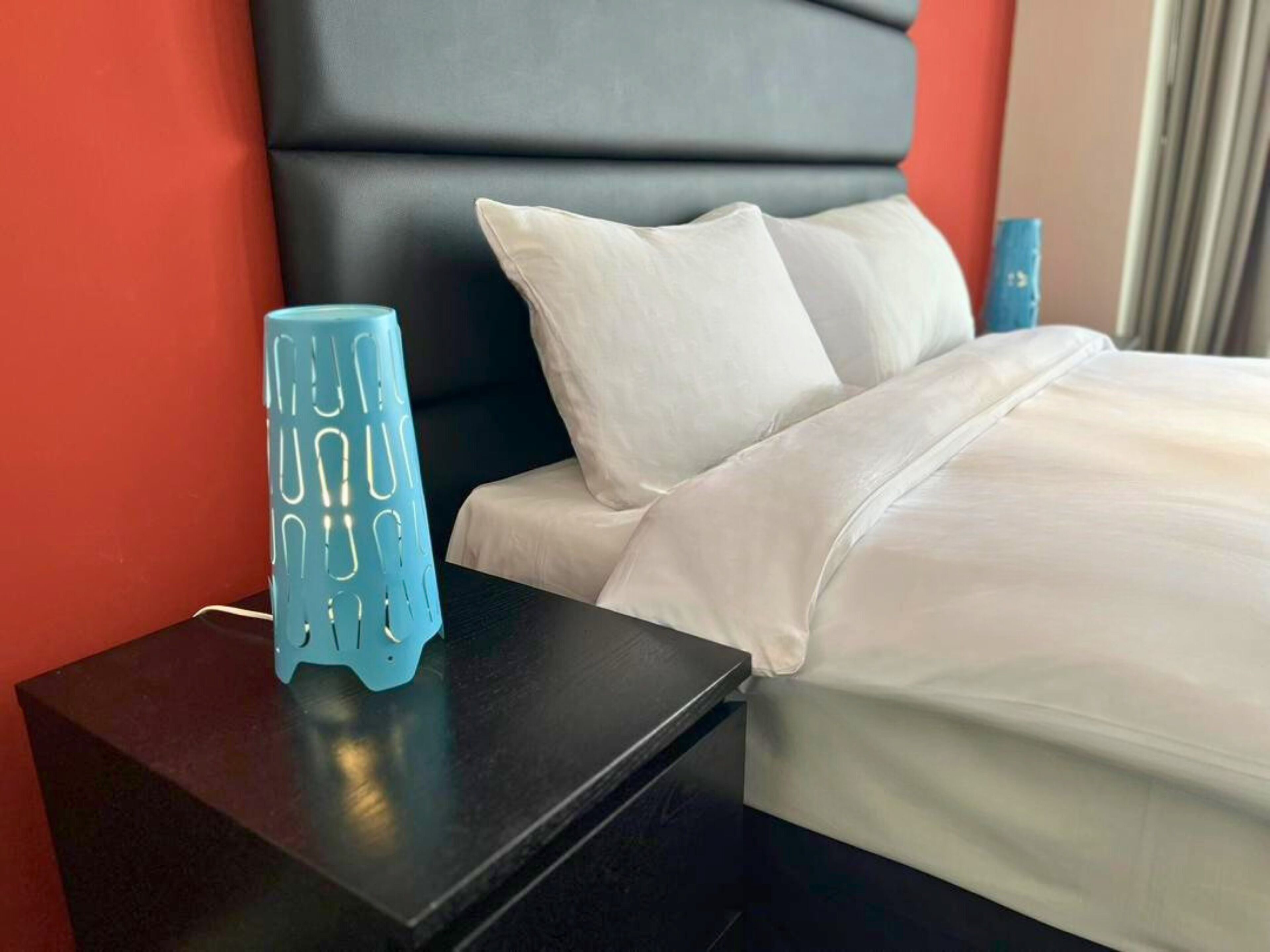 A hotel room with a black nightstand, a white bed with a grey headboard, and a blue vase with a geometric pattern.