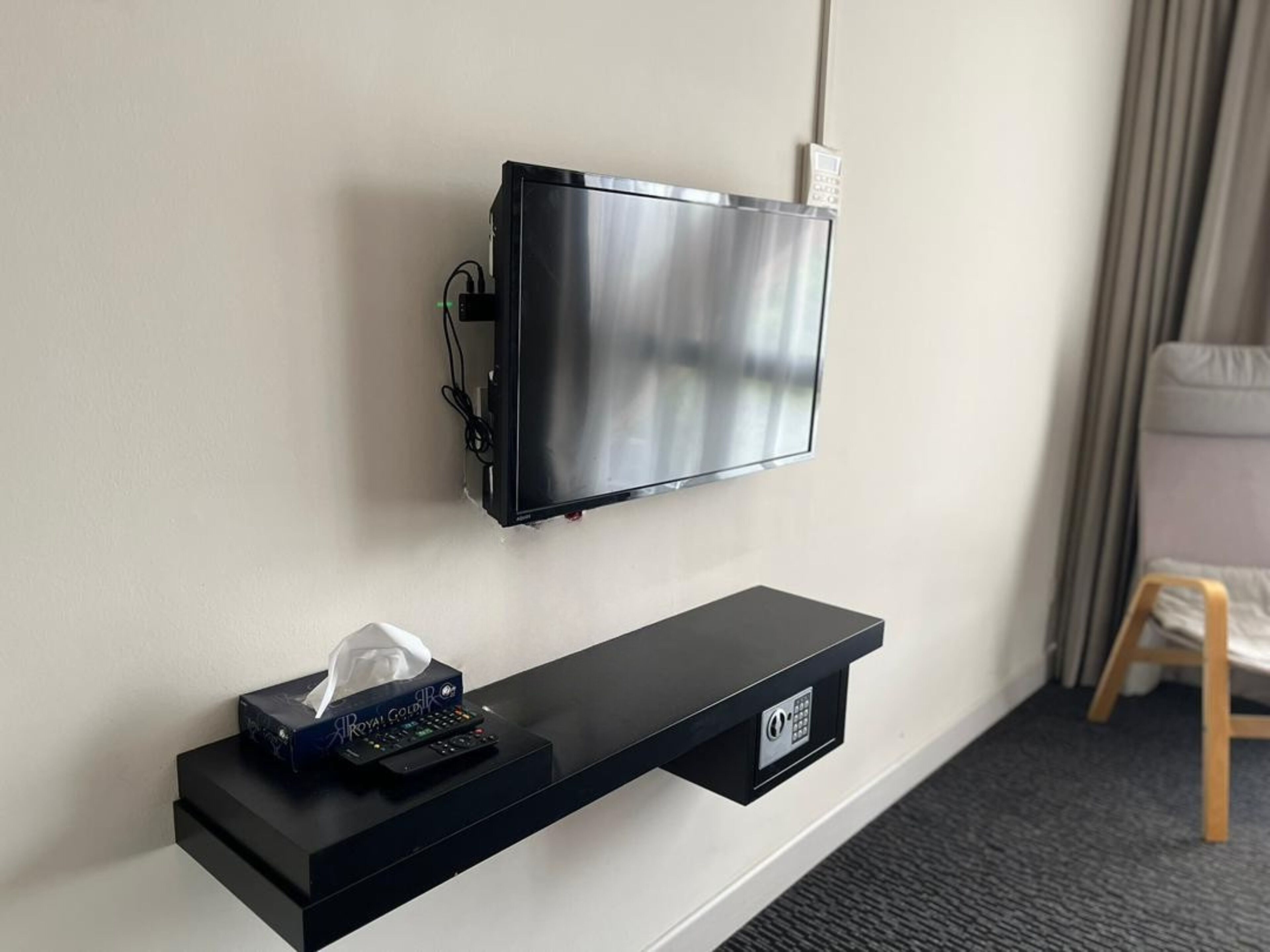 A wall-mounted TV with a remote control and a tissue box on a black shelf below.