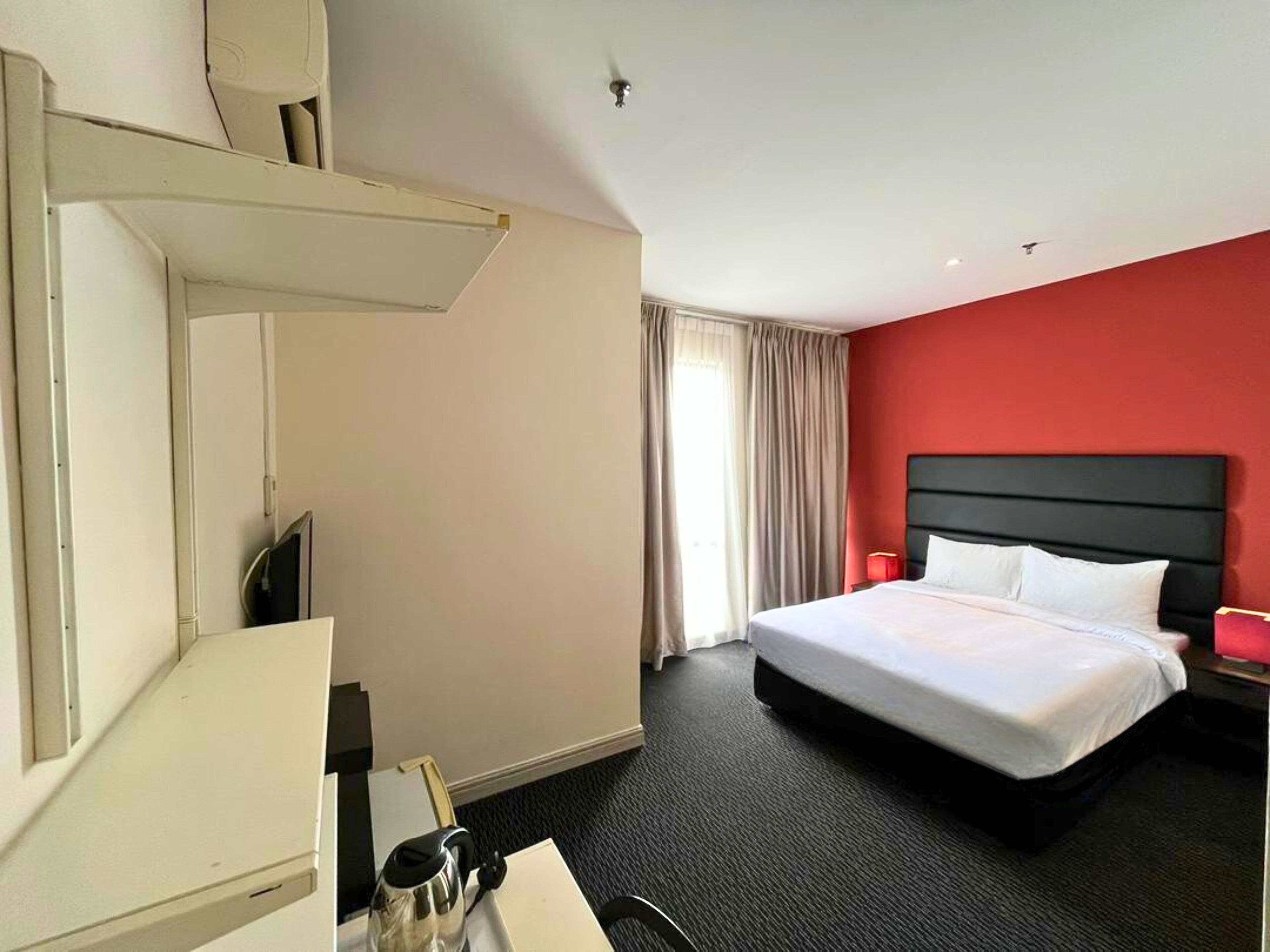 A hotel room with a bed, a desk with a chair, a red lamp, and a window with curtains.