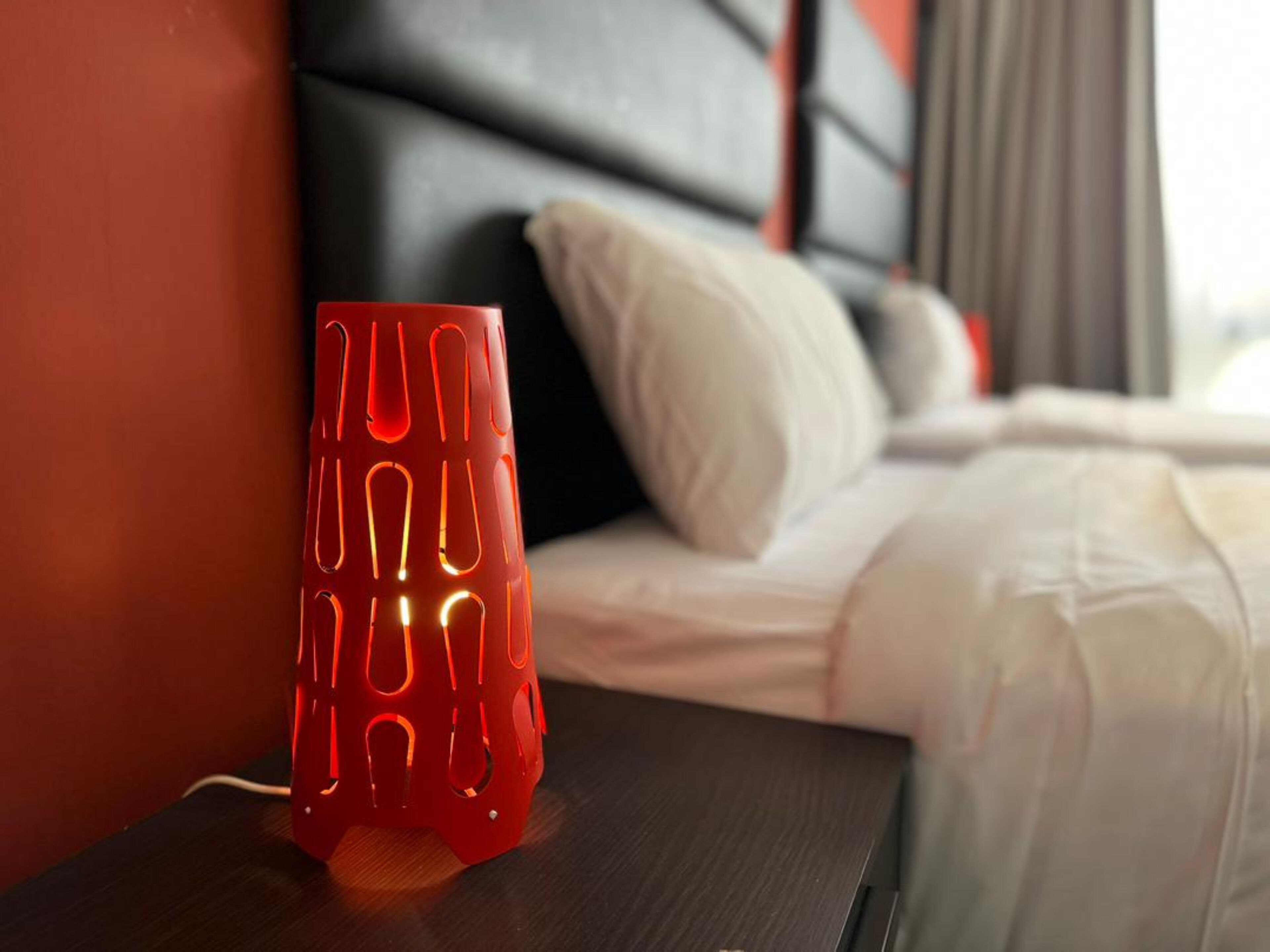 A red candle holder on a bedside table next to a bed with white bedding and a black headboard.