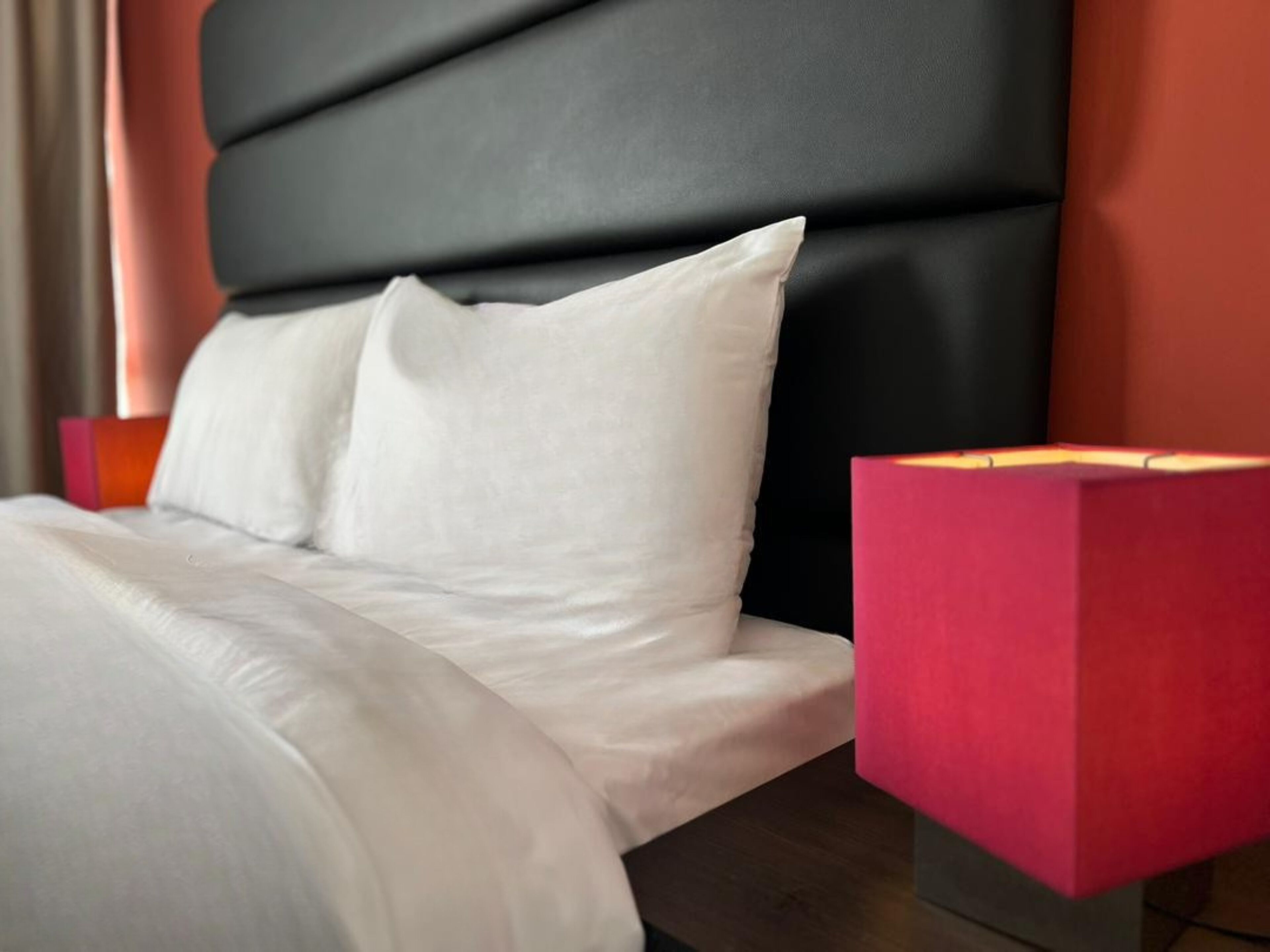 Una cama con ropa de cama blanca y un cabecero negro. Se observa un mesita de noche roja con una lámpara.