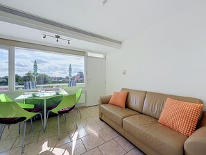 Un salon moderne avec un canapé beige, des fauteuils verts et une table à manger. Une grande fenêtre offre une vue sur les bâtiments et une tour.