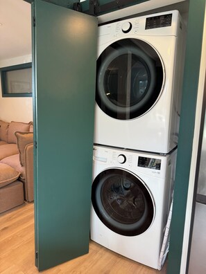 A stacked washer and dryer unit in a room with a wooden floor and a green door. - Spacious Family Apartment w/ off-leash dog park & playground across the street! (Saanich)
