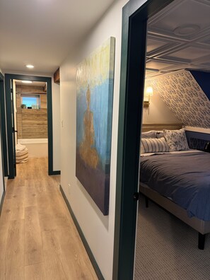 A bedroom with a bed, a painting, a lamp, and a view of a bathroom. - Spacious Family Apartment w/ off-leash dog park & playground across the street! (Saanich)