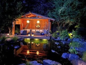 Een houten hutje met een vijver, omringd door rotsen en planten, met buitenmeubels en verlichting.