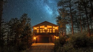 Una cabaña de dos pisos con un garaje amplio, rodeada de árboles y un cielo nocturno estrellado.
