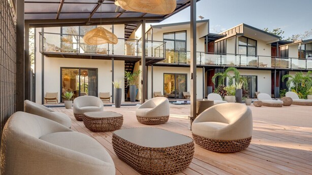 Un espace extérieur moderne, aménagé avec des meubles en osier et doté d’un toit en verre, offrant une vue sur un immeuble à plusieurs étages avec de grandes fenêtres et une piscine.
