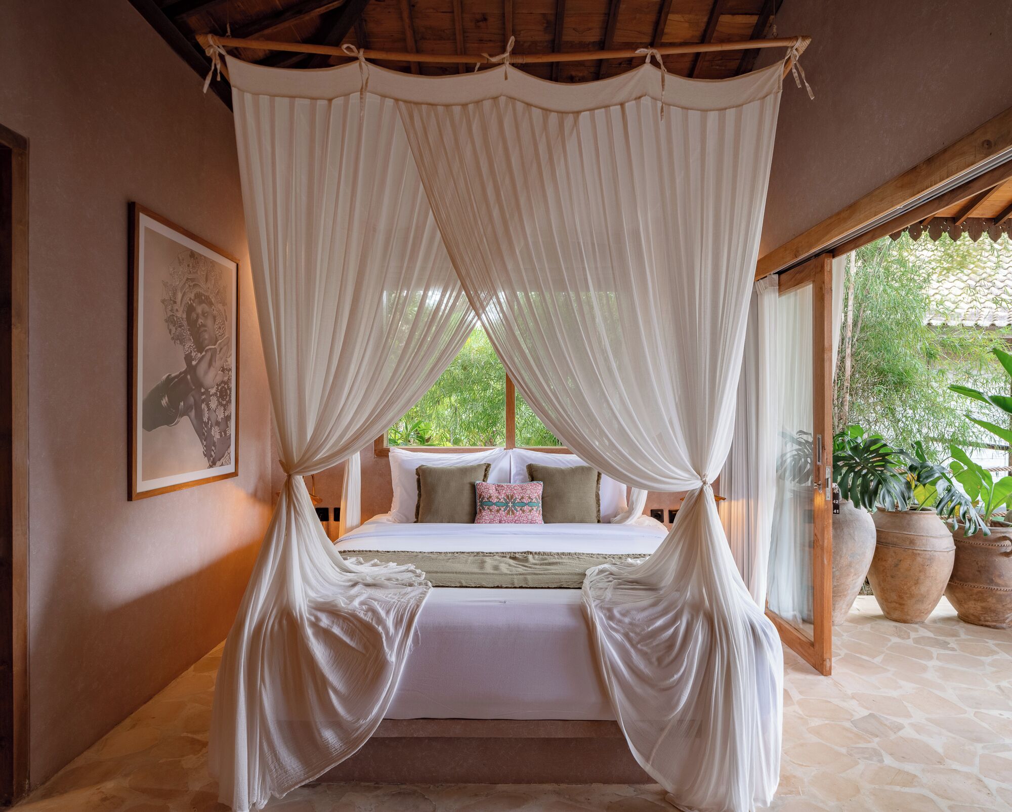 A bedroom with a canopy bed, a framed picture on the wall, and potted plants outside.