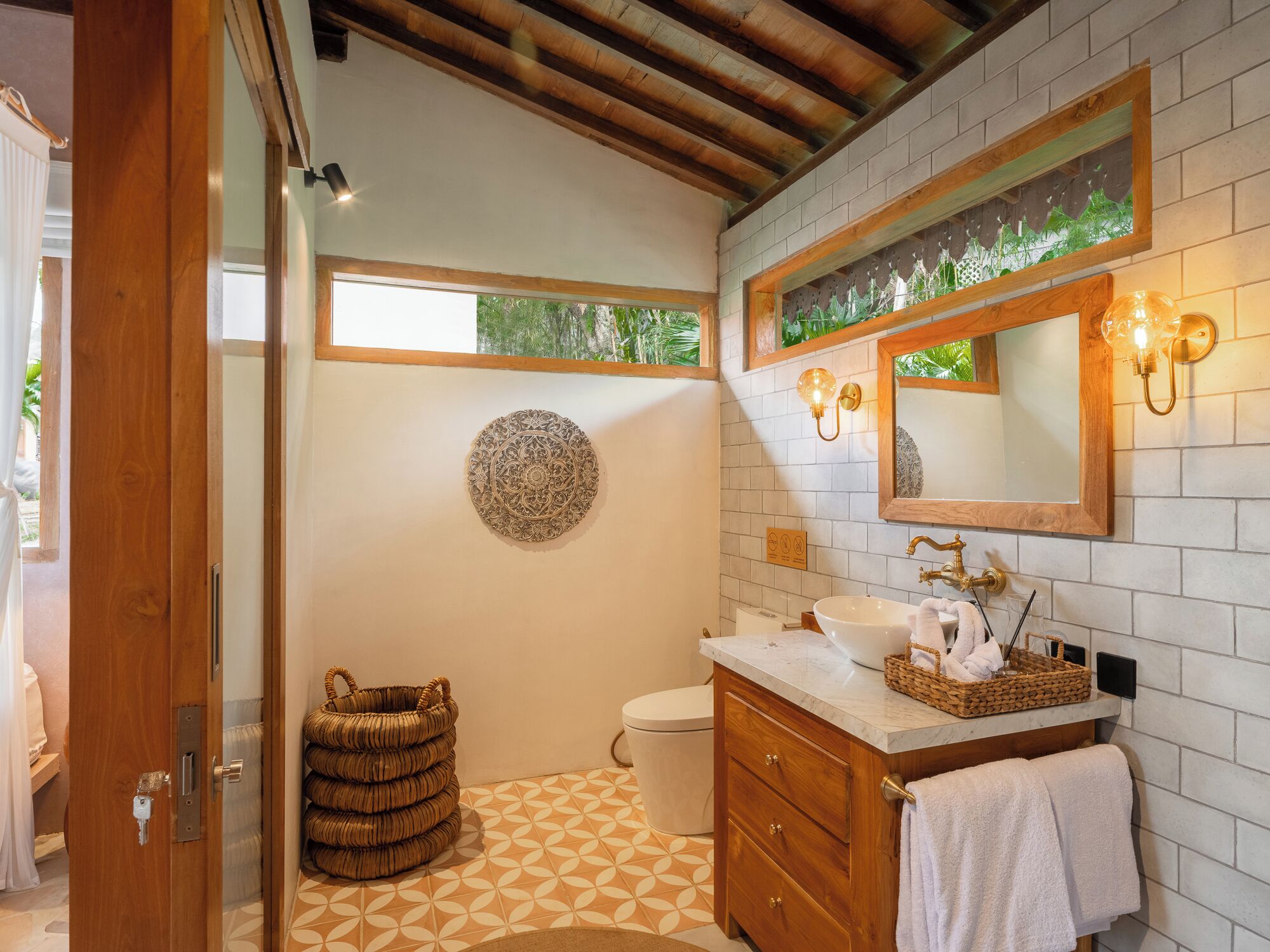Un baño con puerta de madera, un adorno circular en la pared, inodoro, lavabo con encimera de mármol y espejo con apliques de pared.