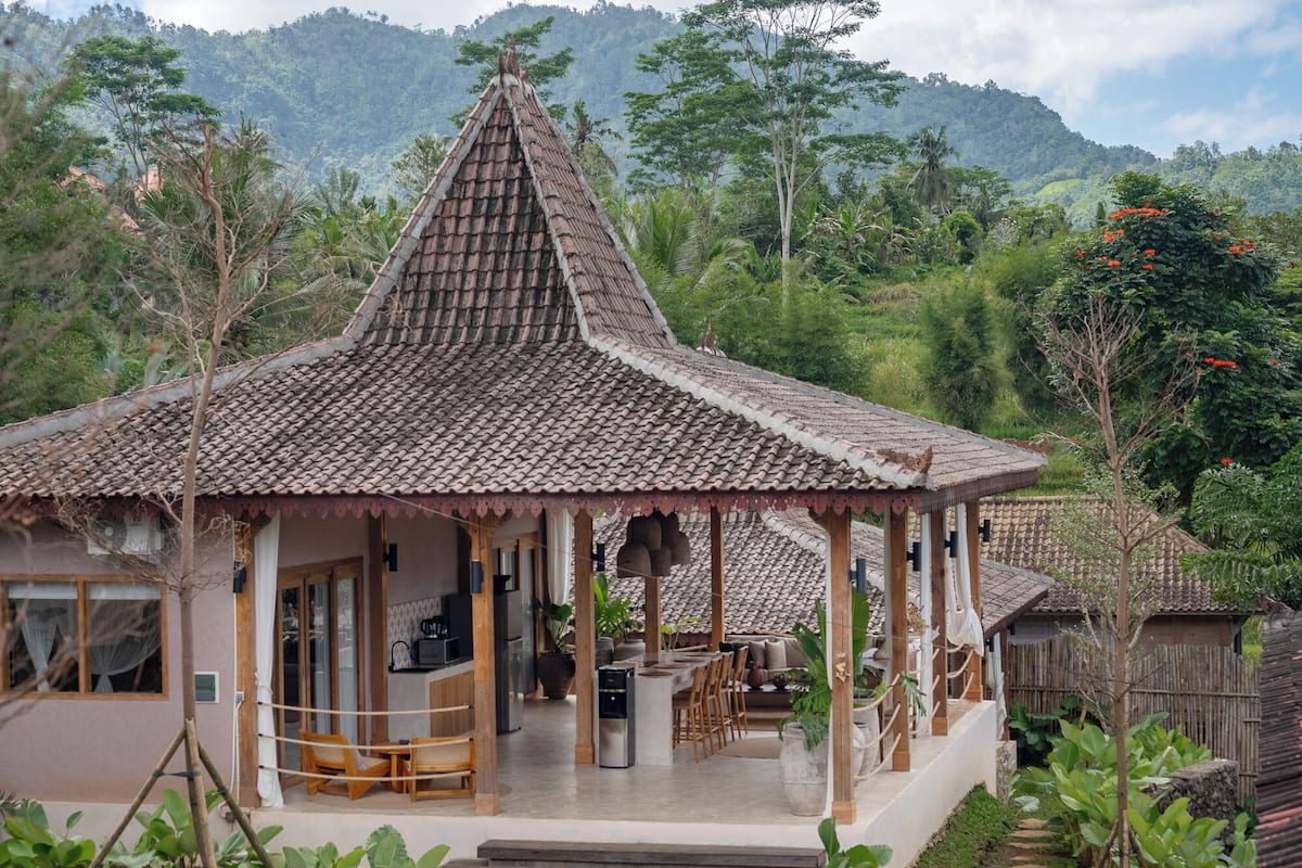 Une maison traditionnelle avec un toit en tuiles, une véranda avec des chaises en bois et un jardin agrémenté d’un chemin de pierres.