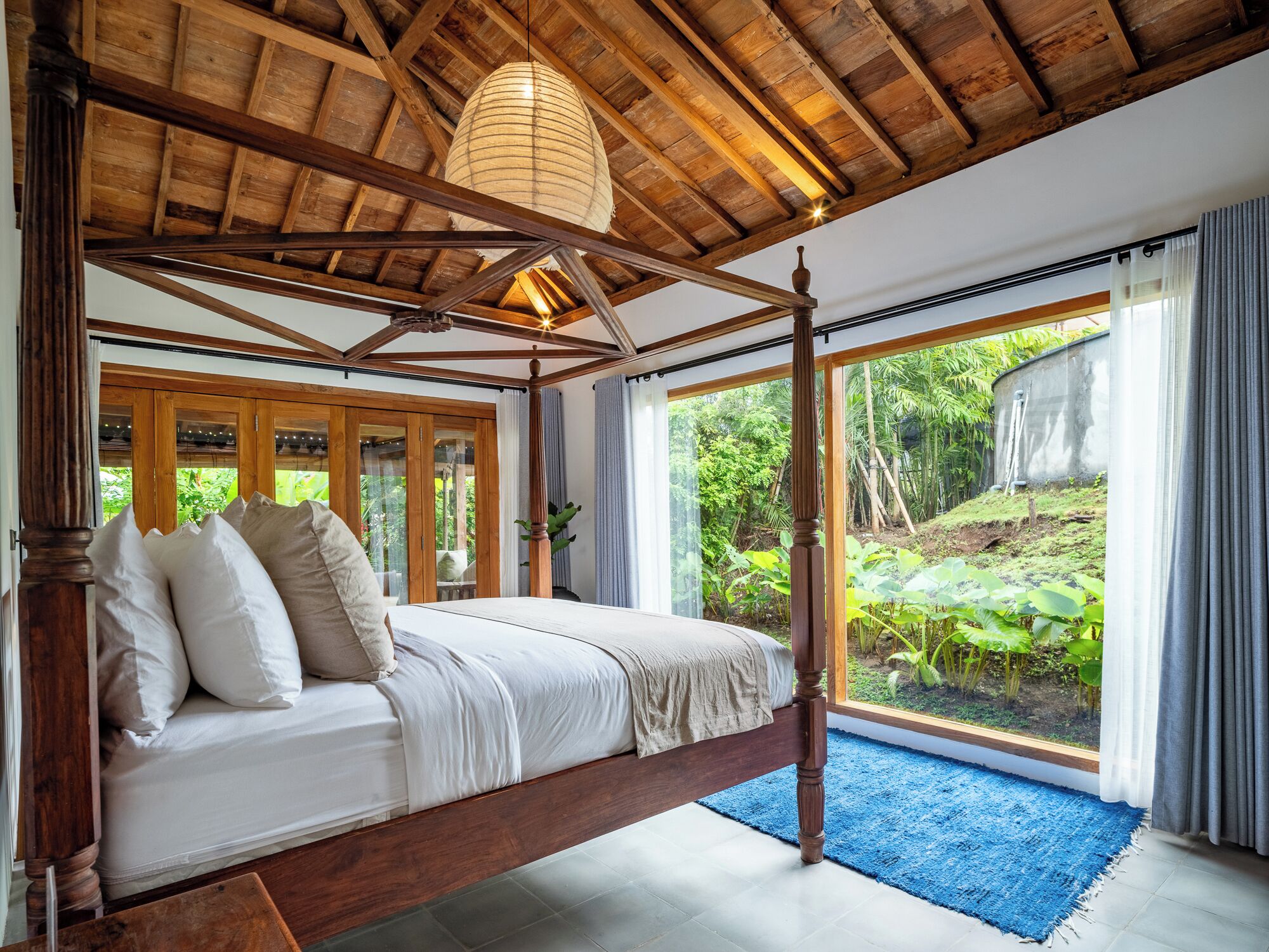A four-poster bed with a wooden frame and white bedding, large windows with curtains, and a view of greenery outside.