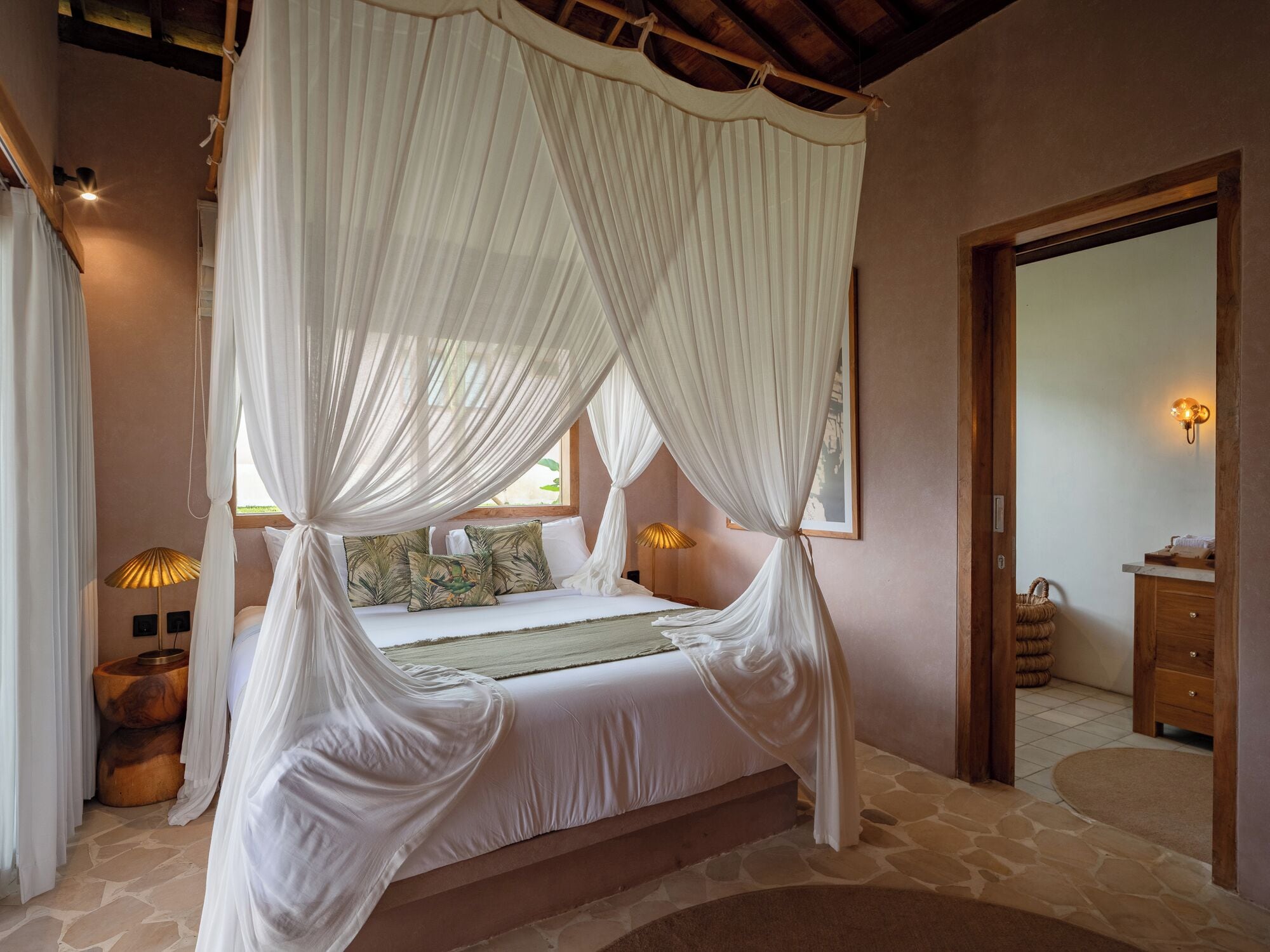 A bedroom with a canopy bed, a wooden headboard, a bedside table with a lamp, and a view of the bathroom.
