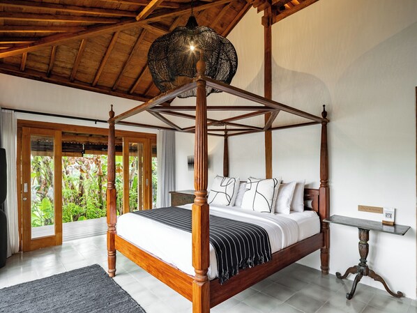 A four-poster bed with a wooden frame and a hanging light fixture, a small table, and a view of greenery through a glass door.