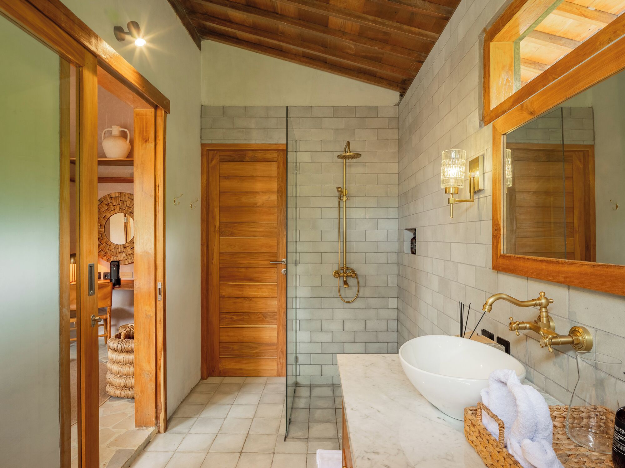A bathroom with a wooden door, a shower with a gold showerhead, a white sink with a large basin, and a mirror with a wooden frame.