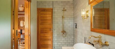 A bathroom with a wooden door, a shower with a gold showerhead, a white sink with a large basin, and a mirror with a wooden frame.