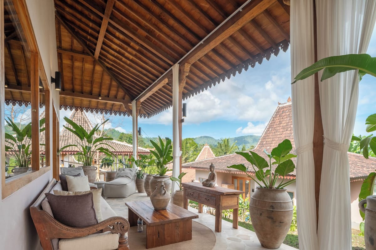 Un espace extérieur aménagé, abrité et doté de mobilier en bois, de grands pots de plantes et offrant une vue sur des bâtiments traditionnels et des montagnes.