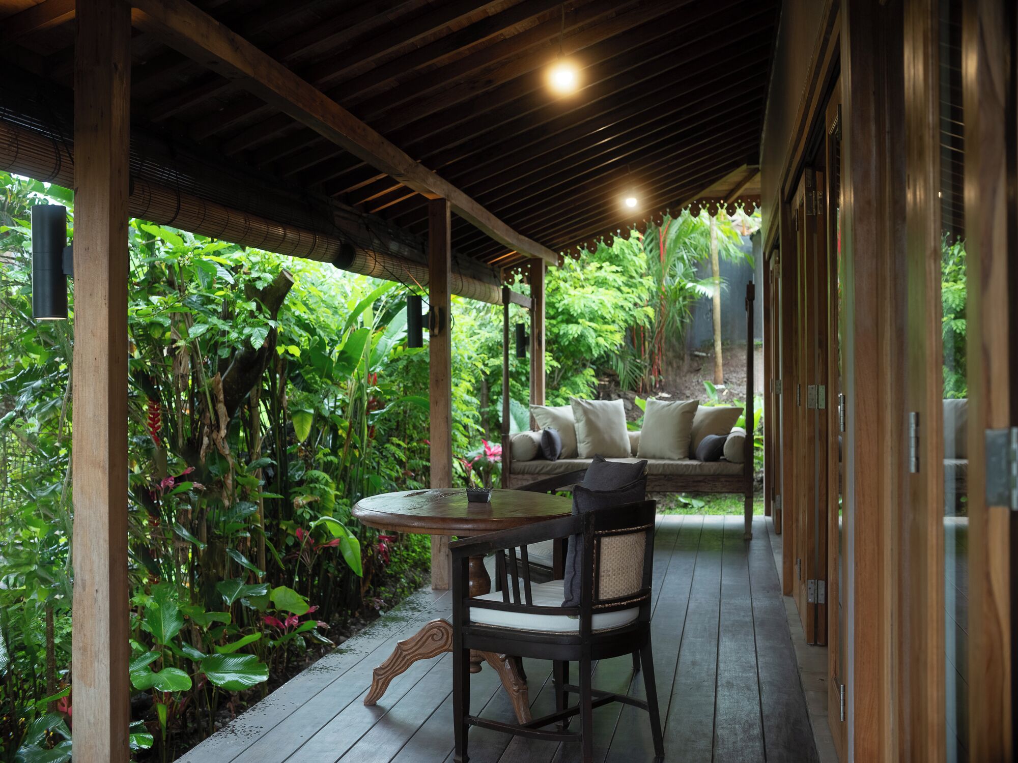 A covered outdoor seating area with a wooden table and chairs, surrounded by lush greenery and a wooden deck.