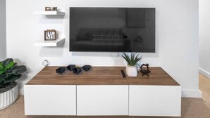 A modern living room with a large flat-screen TV mounted on a white wall, a wooden coffee table, and minimalist decor including a potted plant and decorative items on floating shelves. - Casa Canova | Duplex Full Home For 14PPL @San Juan (San Juan)