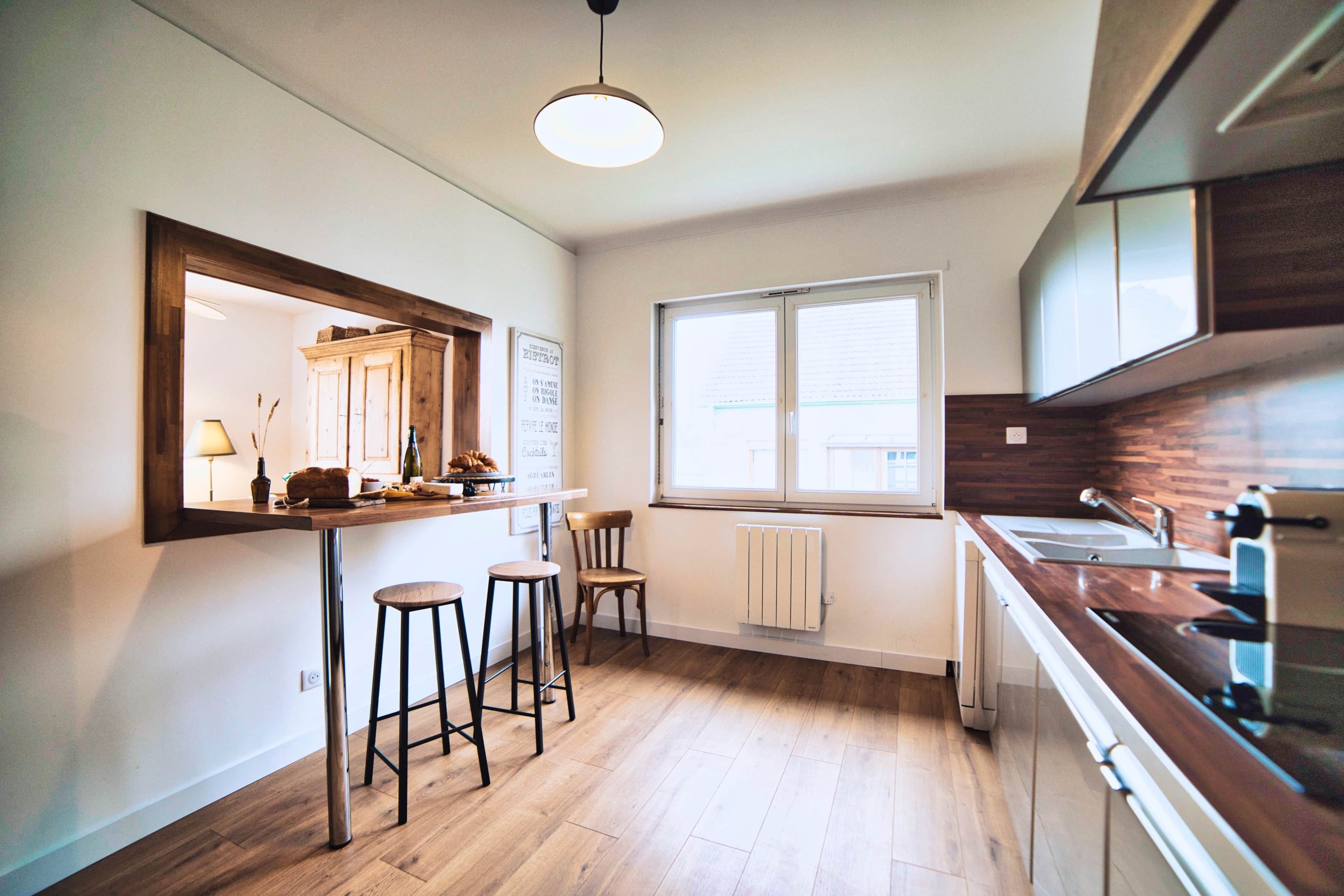 A modern kitchen with wooden flooring, a bar counter with stools, a large window, and built-in appliances.