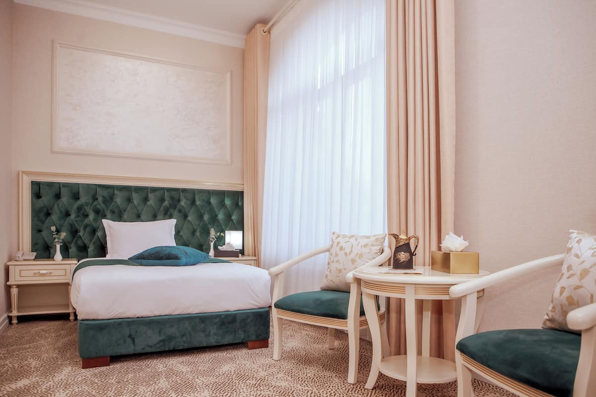 A hotel room with a large bed, a green upholstered headboard, a small table with a vase and tissues, and a chair with a green cushion.