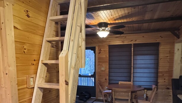 Un interior de madera con una escalera de madera, una zona de comedor con una mesa y sillas, y un ventilador de techo.