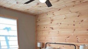 Un dormitorio con techo de madera, una cama con estructura metálica, dos mesitas de noche con lámparas y una ventana con persianas.