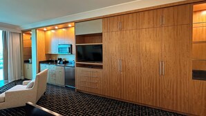 A modern hotel room with a large bed, a seating area, a kitchenette, and wooden cabinets.