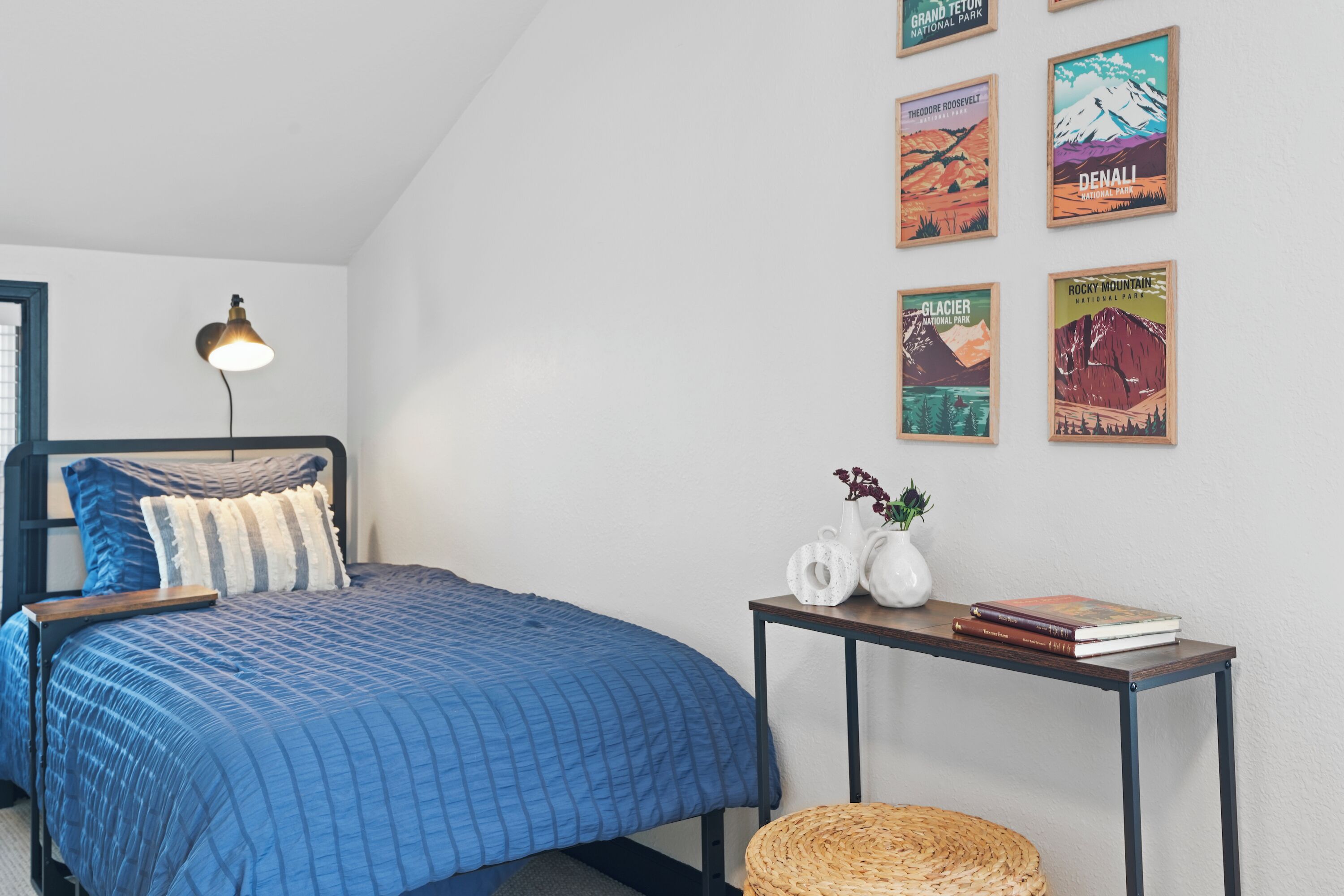 Una camera da letto con un letto, una lampada da comodino, un tavolino con libri e un vaso, e una parete decorata con poster incorniciati.