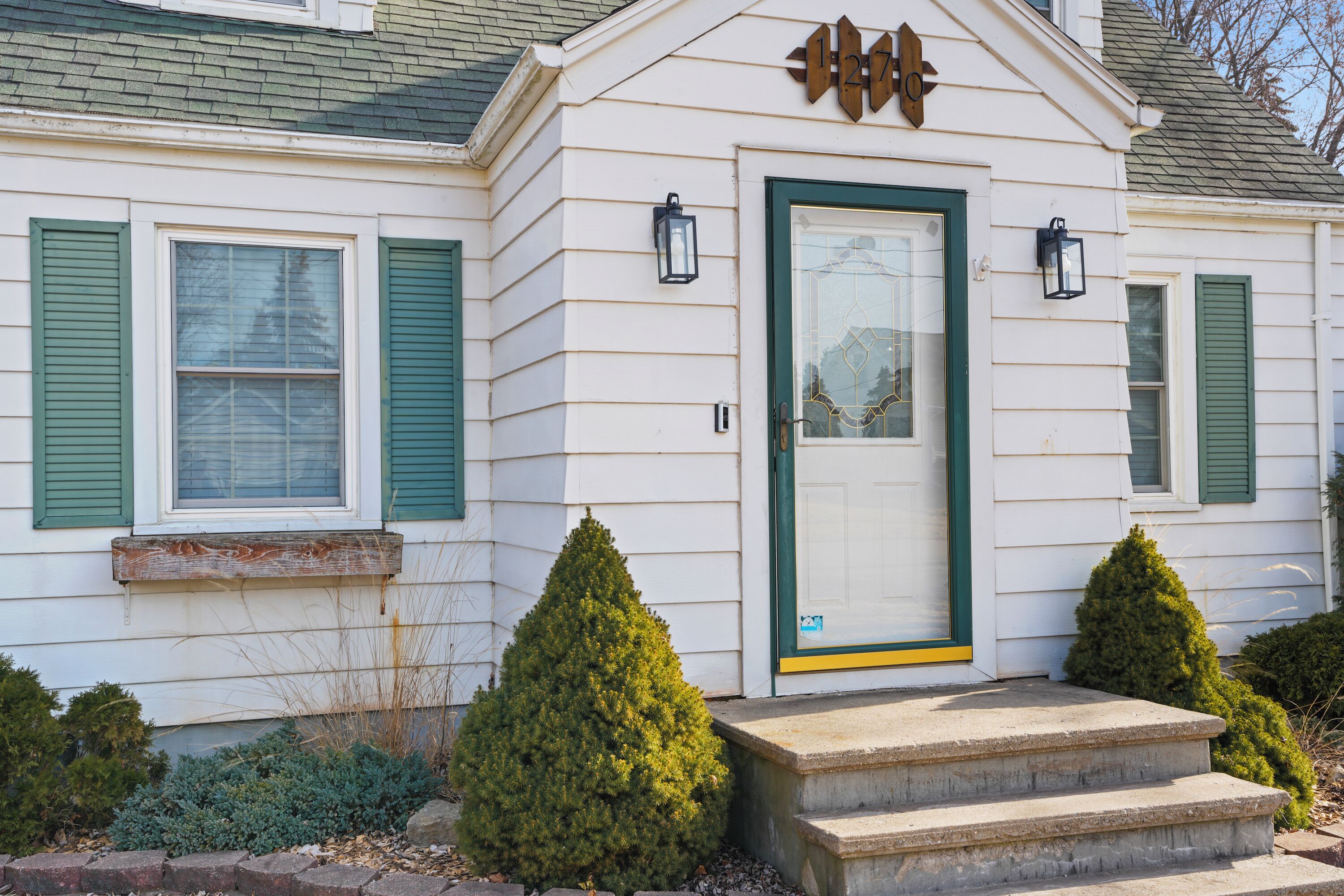 A white house with green shutters, a front door with a green frame, and two small trees in front.