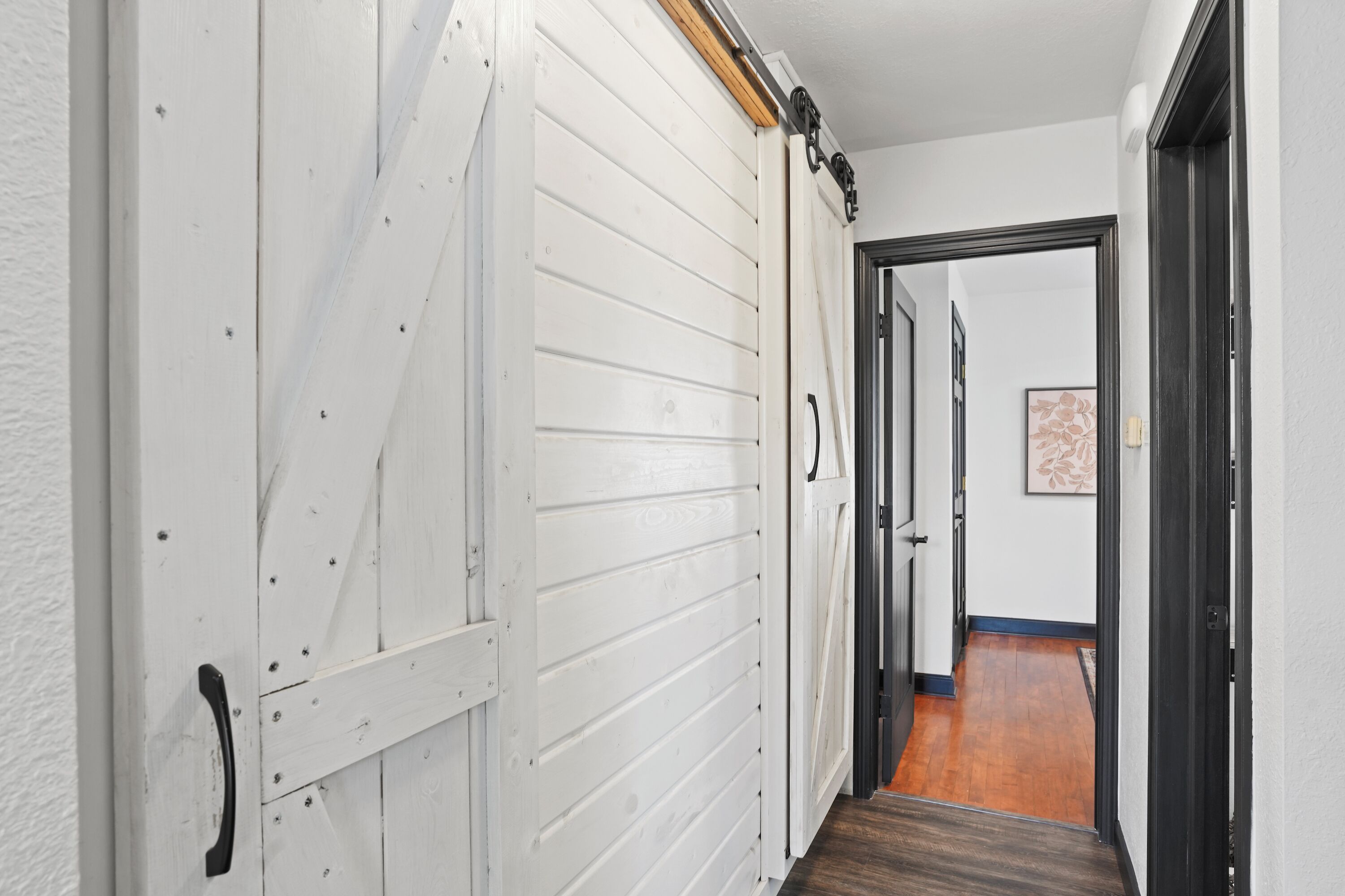 A hallway with a white door and a black door leading to a room with a wooden floor.