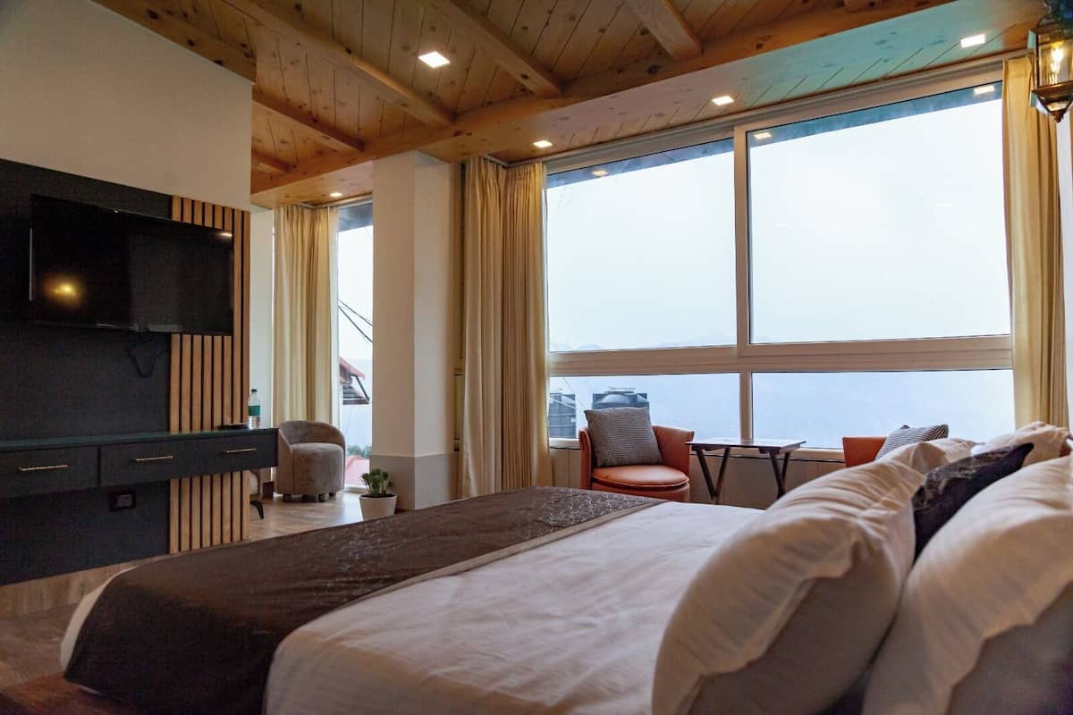 Une chambre d’hôtel avec un grand lit, un téléviseur fixé au mur, une petite table et une chaise. Il y a une grande fenêtre avec des rideaux, et la vue donne sur l’extérieur.