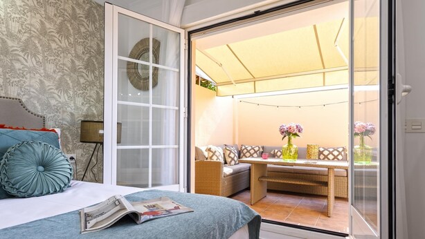 Une chambre à coucher comprenant un lit, une table de chevet, une lampe et une porte-fenêtre coulissante donnant sur un balcon aménagé avec des meubles en osier et une table.