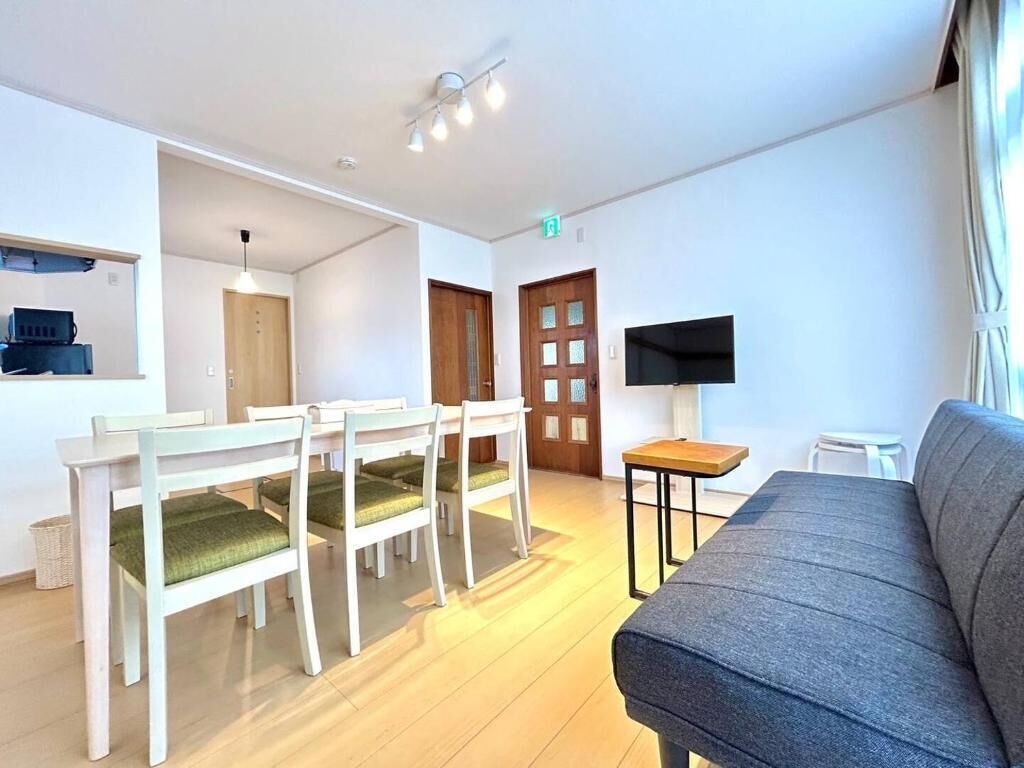 A living room with a grey sofa, a dining table with chairs, a TV on a stand, and a wooden door.