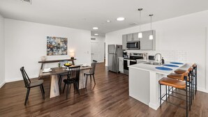 Eine moderne Küche mit Edelstahlgeräten, ein Essbereich mit einem Holztisch und -stühlen sowie eine weiße, geflieste Spritzschutzwand.