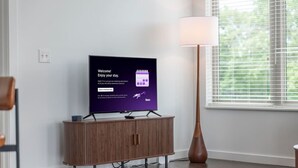 Ein modernes Wohnzimmer mit einem Flachbildfernseher auf einem Holzständer, einer Stehlampe und einem Fenster mit Jalousien.