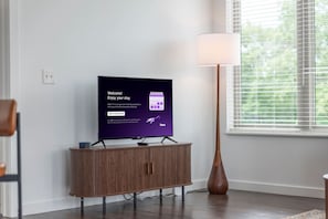 Moderni olohuone, jossa on litteänäyttöinen televisio puisen jalustan päällä, lattiavalaisin ja ikkuna, jossa on sälekaihtimet.