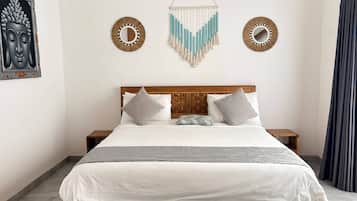 A bedroom with a bed, two nightstands, a wooden headboard, a wall hanging, and a window with curtains.