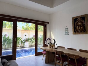 A dining area with a wooden table and chairs, a Buddha statue, and a view of a swimming pool through glass doors. - Amy Guesthouse Nusa Dua (Nusa Dua)