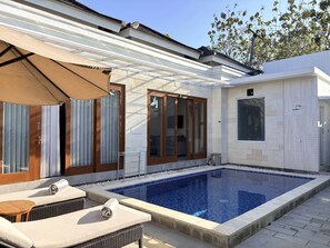 Zona de piscina con sillones de descanso, un espacio de estar techado y un edificio con grandes ventanales y puerta.