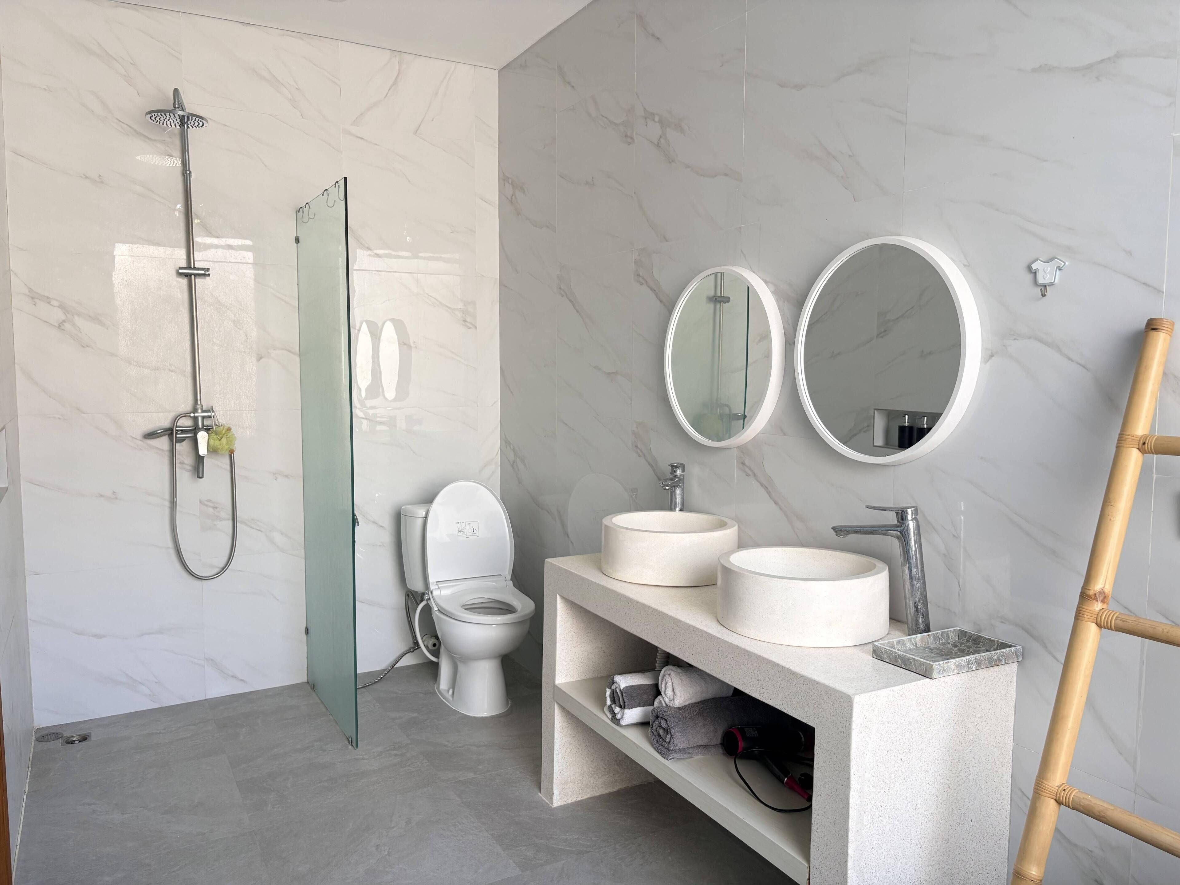 Une salle de bain moderne dotée d’une douche à parois de verre, de toilettes, de deux miroirs ronds et d’un meuble-lavabo avec un évier.
