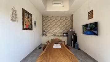 A dining area with a long wooden table, chairs, a kitchen in the background, and a television on the wall.