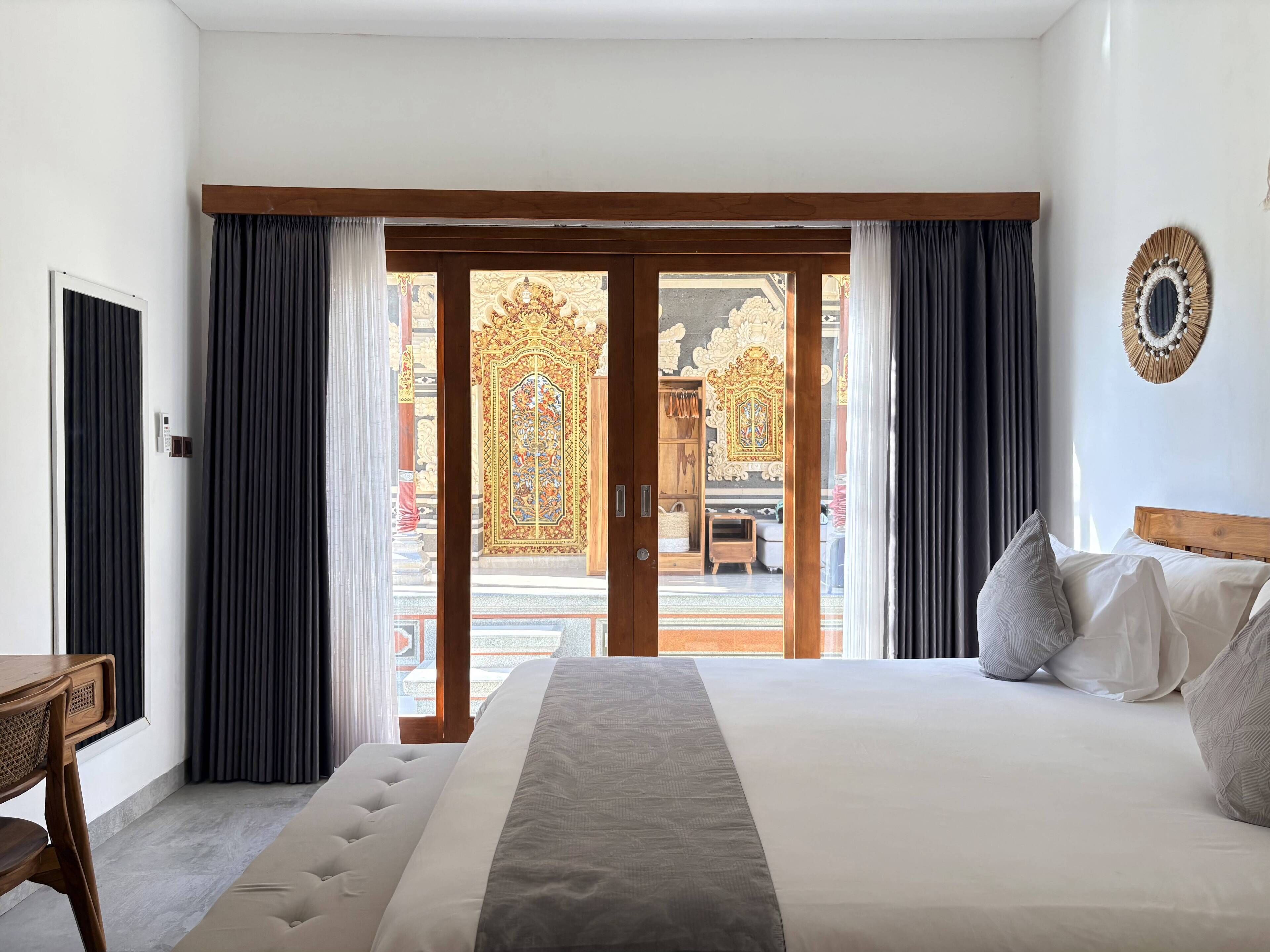 A hotel room with a large bed, a wooden headboard, a desk, and a view of an ornate gate through the sliding doors.