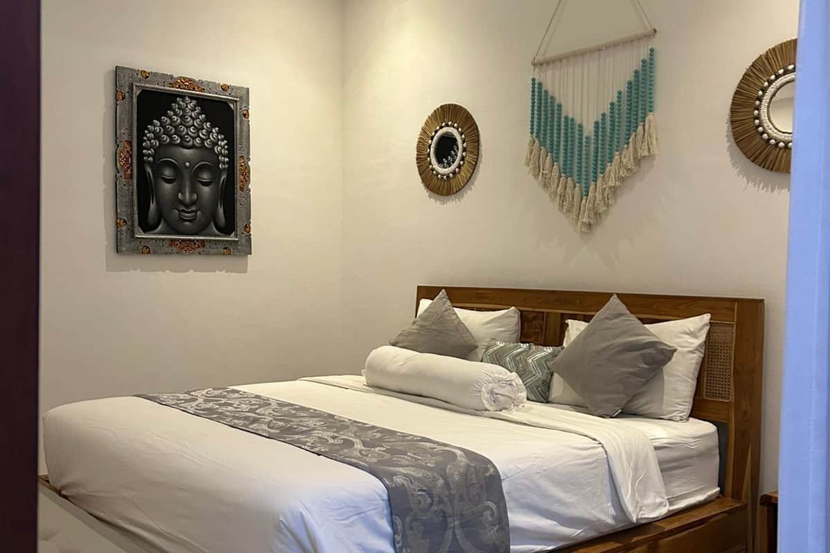 Ein ordentlich bezogenes Bett mit hölzernem Kopfteil, einer Bank am Fußende und Wanddekorationen, darunter ein Buddha-Kopf und zwei runde Spiegel.