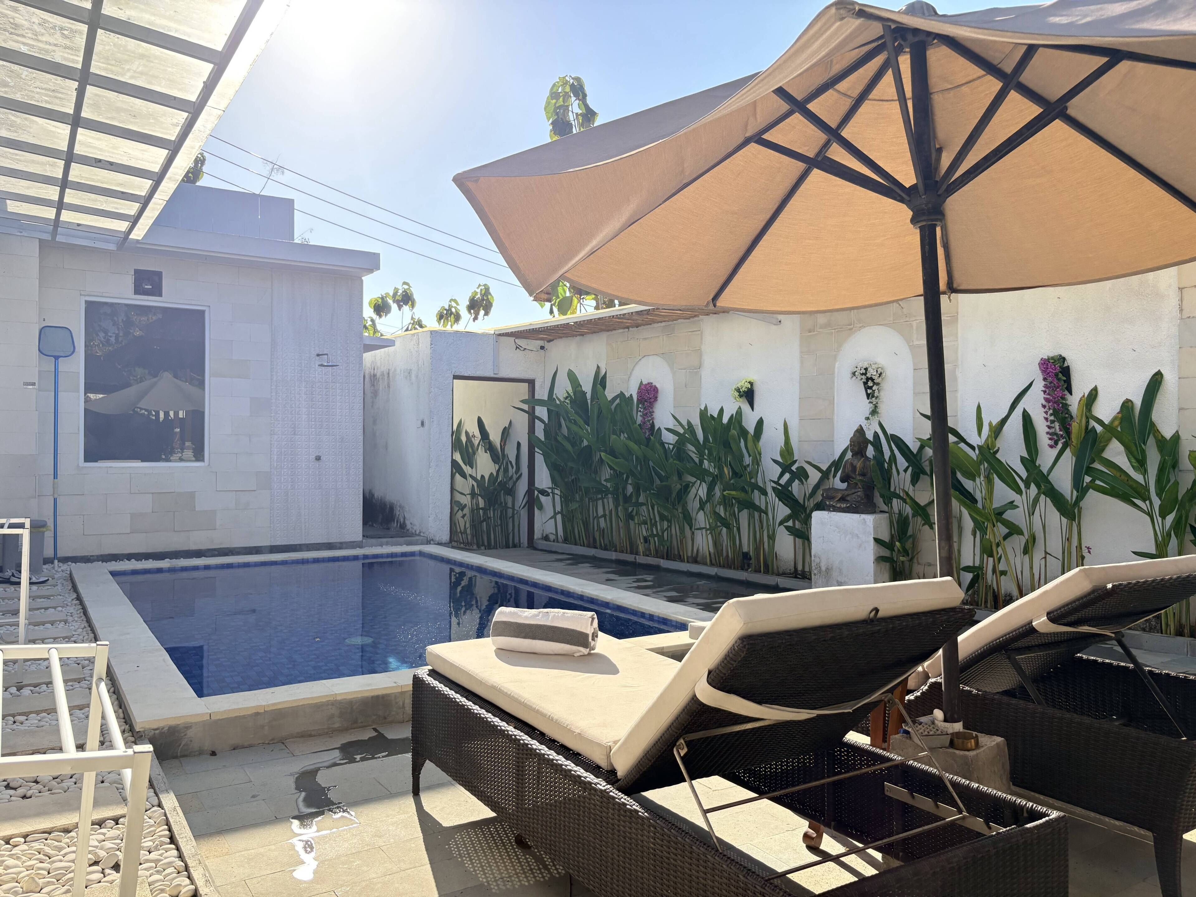A swimming pool with lounge chairs and an umbrella, surrounded by plants and a Buddha statue.