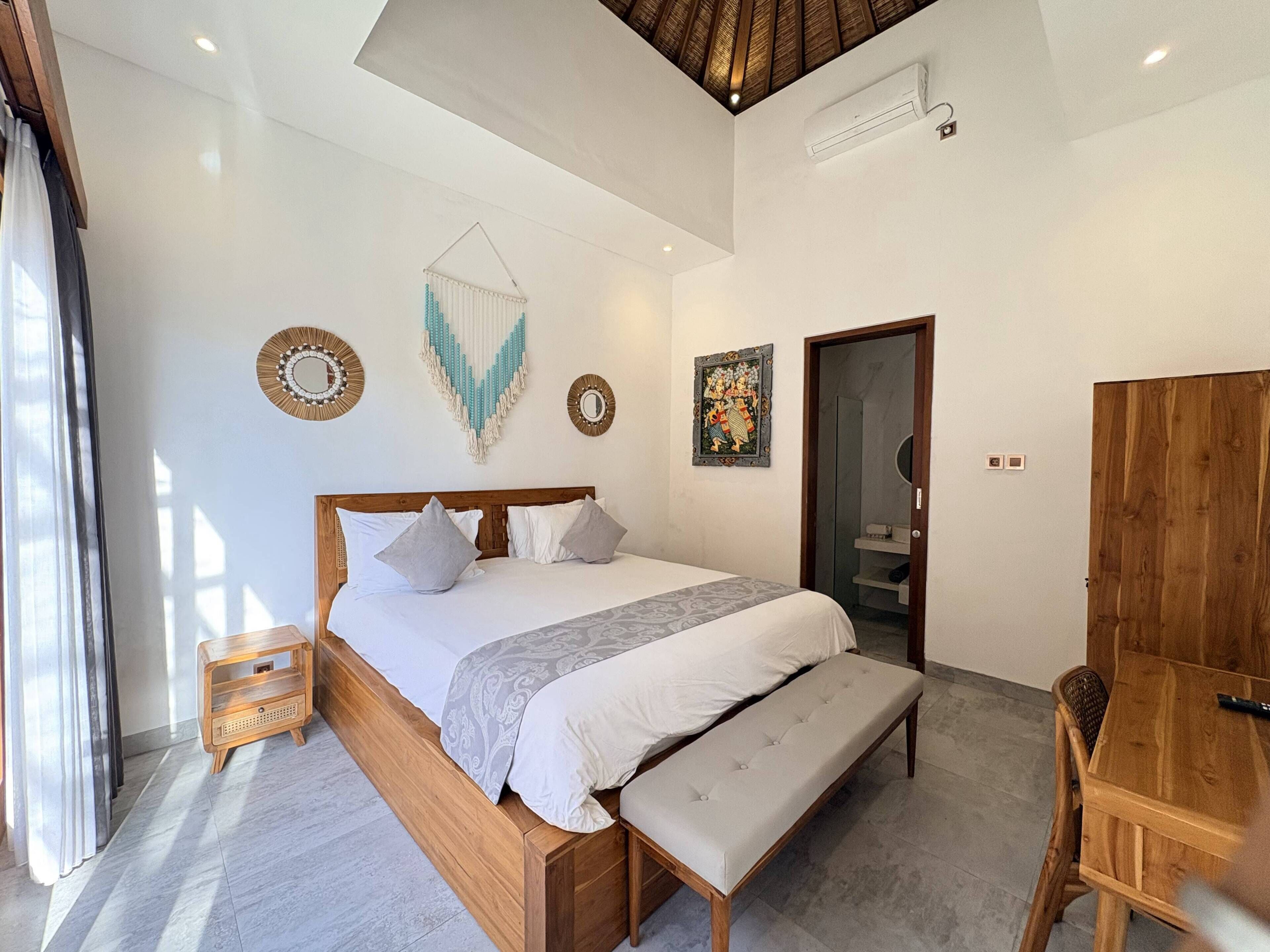 A bedroom with a wooden bed, a bench, a wooden desk, and a door leading to a bathroom.