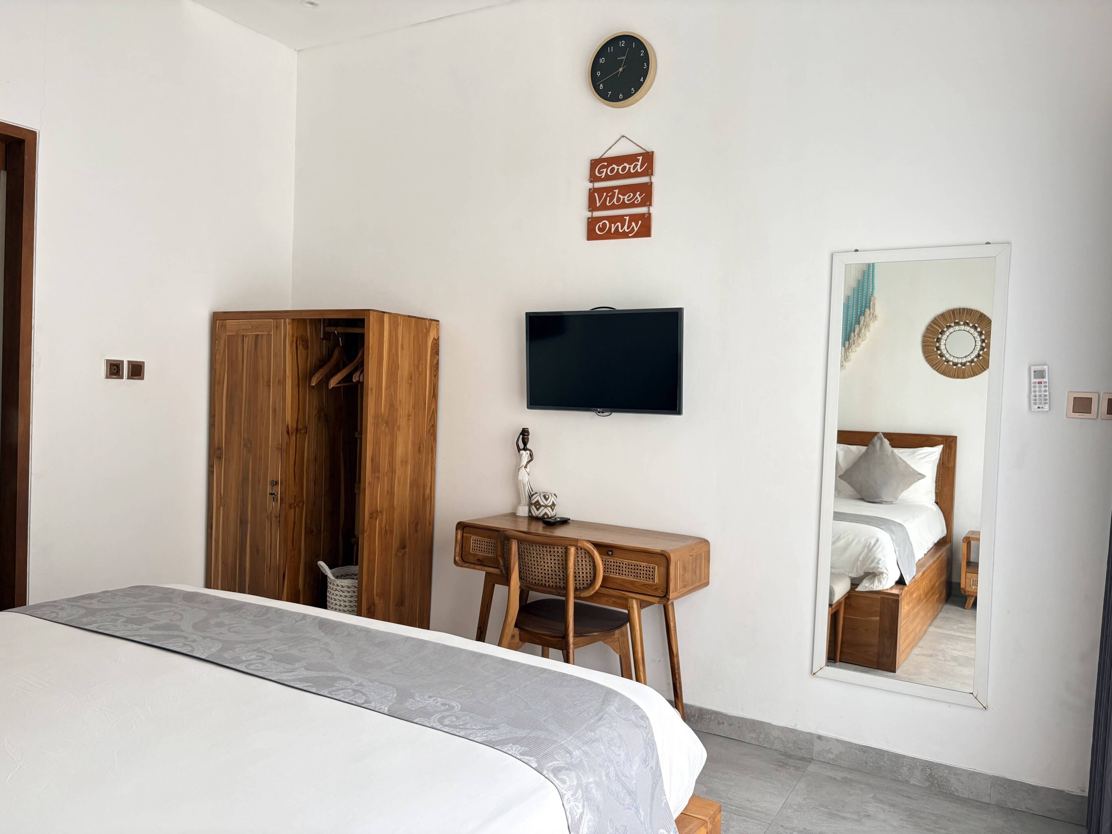 A bedroom with a bed, a wooden wardrobe, a desk with a chair, a television, and a mirror reflecting the bed.
