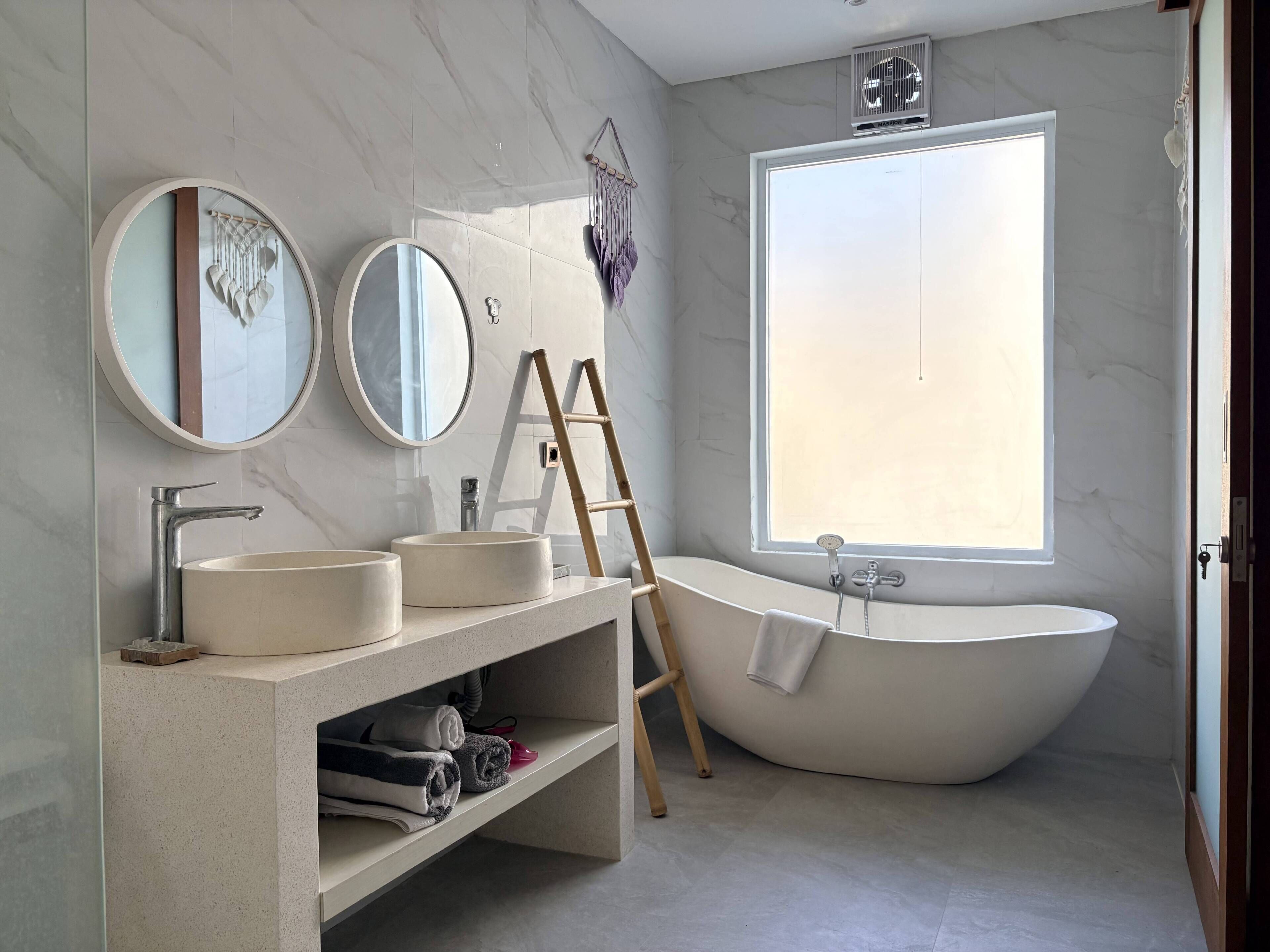 Ein modernes Badezimmer mit freistehender Badewanne, zwei runden Spiegeln, einem Waschtisch und einem Fenster für natürliches Licht.