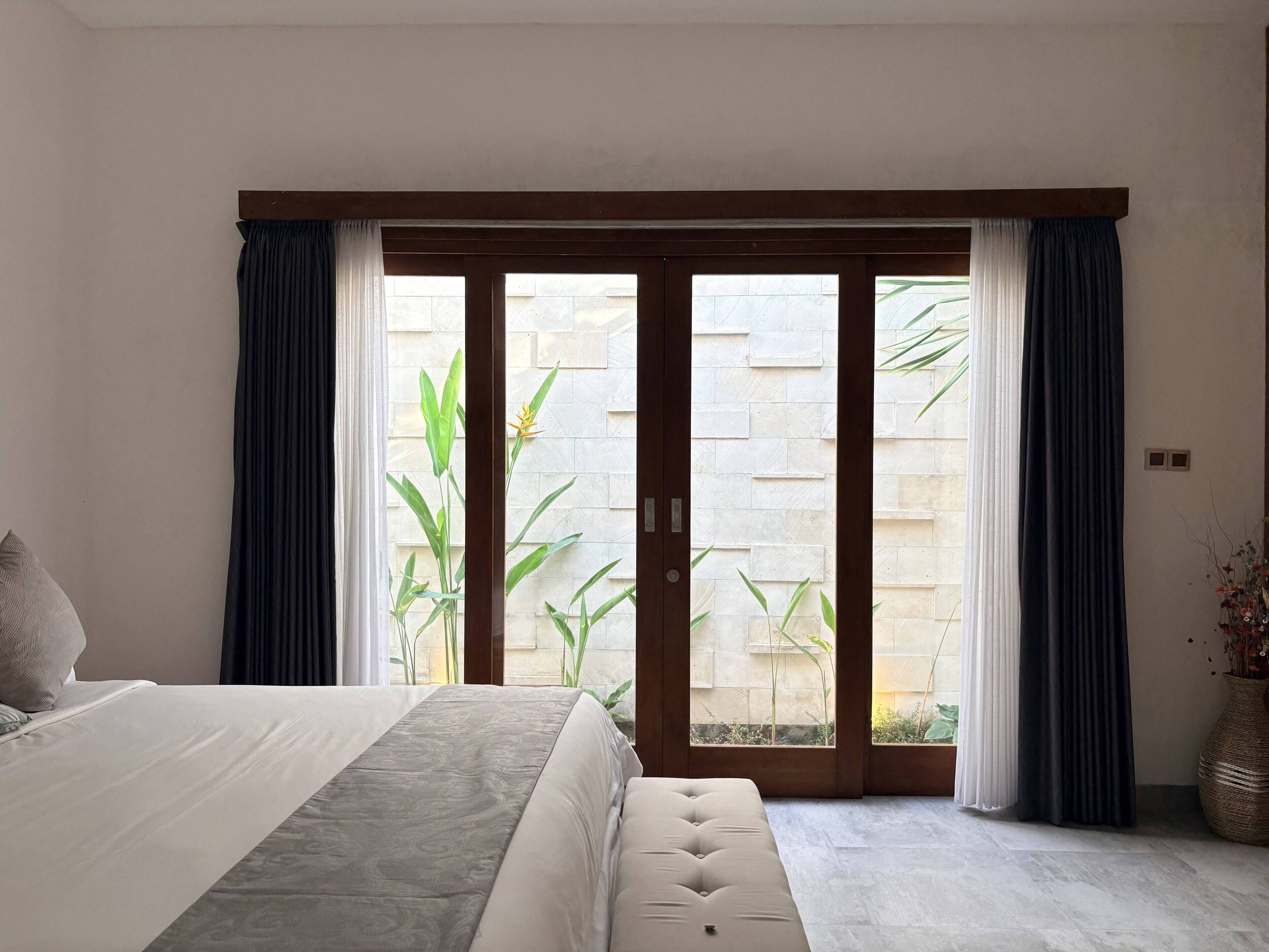 A bedroom with a bed, a bench, a sliding glass door to an outdoor area, and a potted plant.
