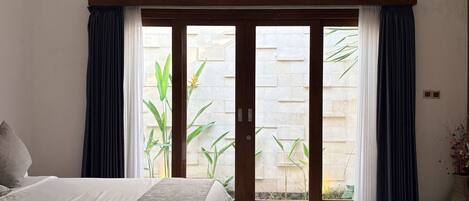 A bedroom with a bed, a bench, a sliding glass door to an outdoor area, and a potted plant.