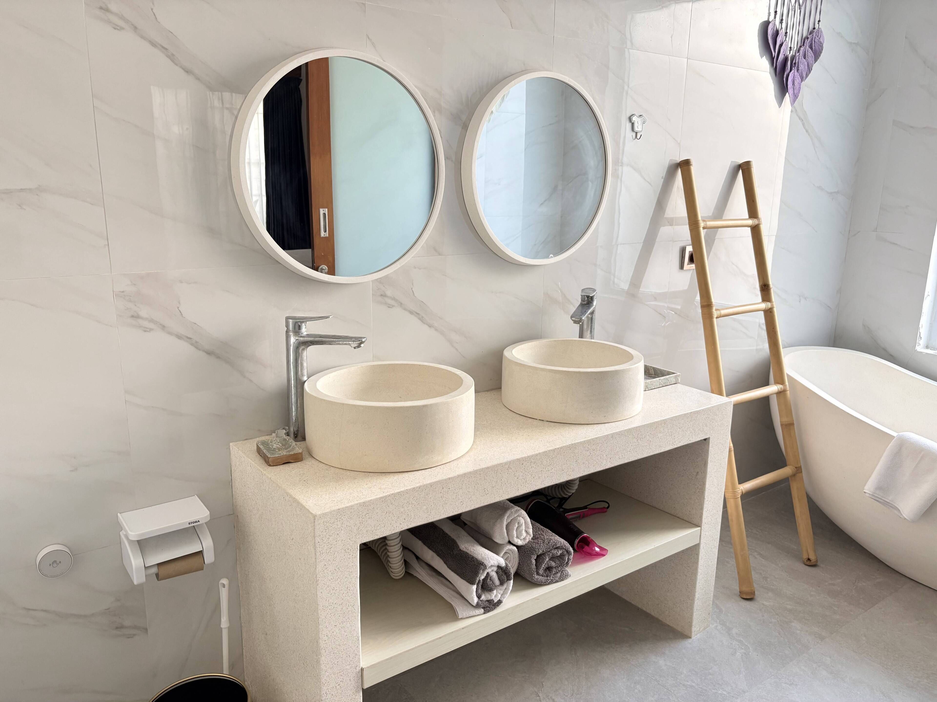A bathroom with two round sinks, a mirror, a towel rack, a ladder, and a toilet paper dispenser.